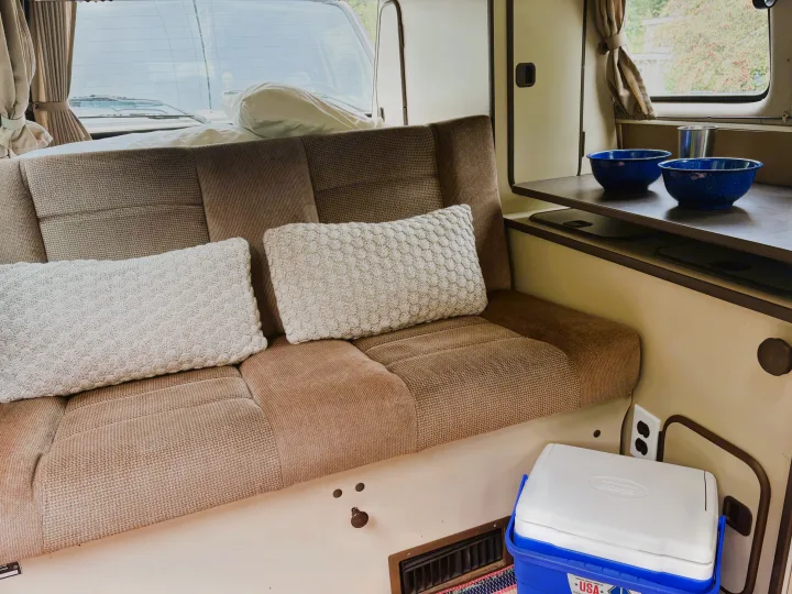 Cozy rv rental interior: A comfortable bench seat, pillows, dishes on a table, and a cooler. Ready for van life adventures!. Volkswagen Westfalia 1985