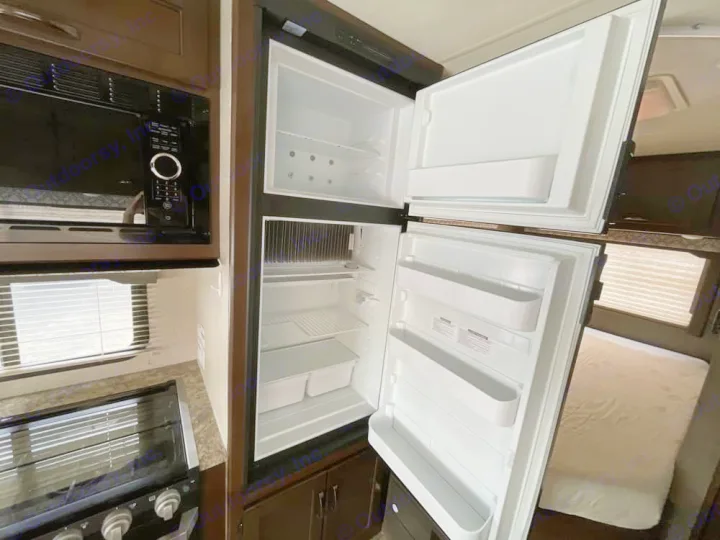 An RV rental kitchen features an open top-freezer refrigerator, a microwave, and stovetop. The fridge interior shows shelves, and a window is visible.. Thor Freedom Elite 2020