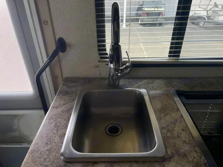 RV rental kitchen: A stainless steel sink with a modern faucet, countertop, and window with blinds overlooking a parked vehicle.. Thor Freedom Elite 2020