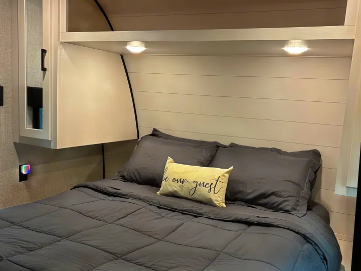 This RV rental offers a cozy bedroom with a dark gray bed, decorative pillow, shiplap-style headboard, overhead storage, and ambient lighting.. Jayco Jay Flight SLX 2024