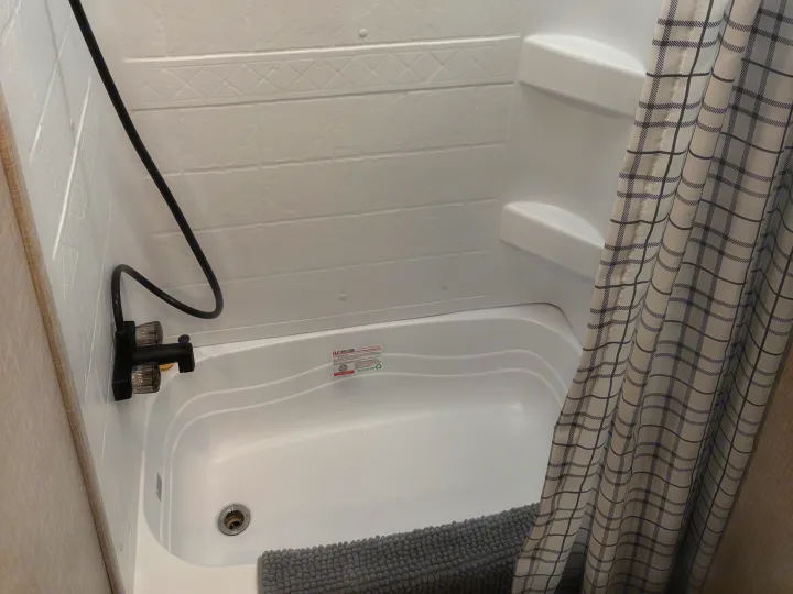 Inside an RV rental: a modern white shower/tub combo with a black faucet, plaid curtain, and gray bath mat.. Jayco Jay Flight SLX 2024