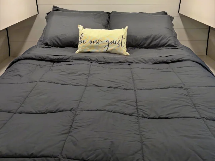 An inviting, neatly made bed in an RV rental, featuring dark gray bedding and a "be our guest" pillow.. Jayco Jay Flight SLX 2024