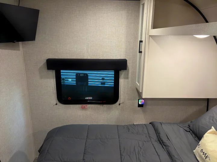 Inside a compact RV rental bedroom: grey bed, TV, window with 'EXIT' sign, overhead storage & colorful nightlight.. Jayco Jay Flight SLX 2024