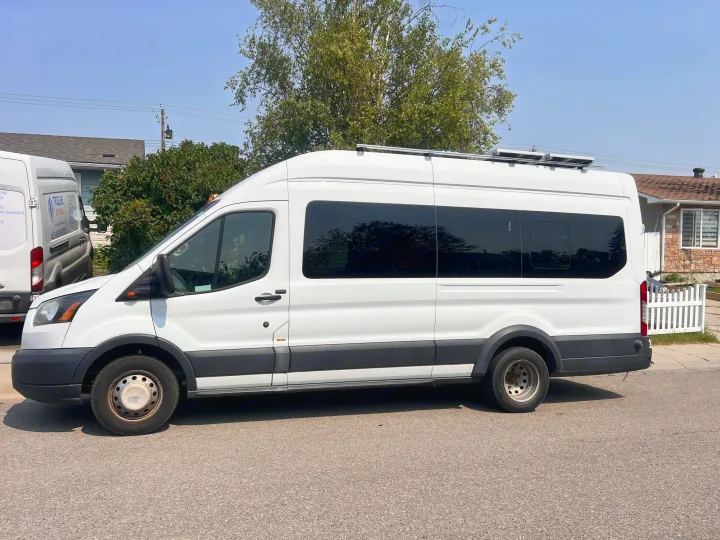 White high-roof Ford Transit van with tinted windows & solar panels on roof, suggesting an RV rental conversion, parked outdoors.. Ford Transit 2016