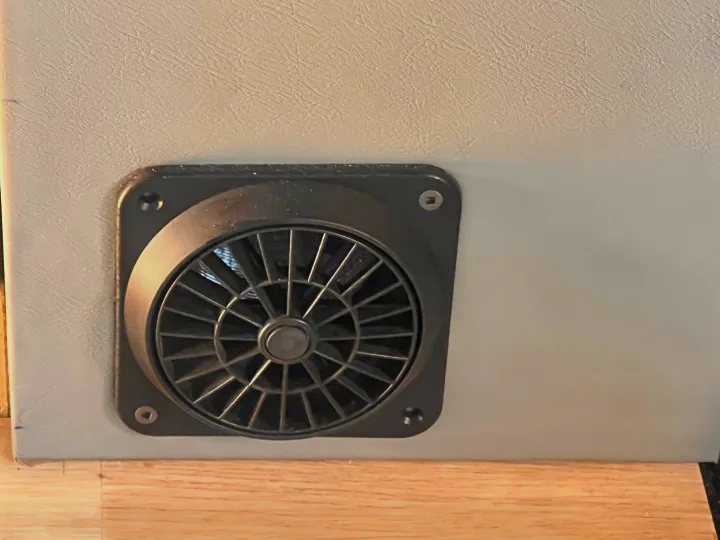 Air circulation vent in an RV rental. A black square vent with a circular grille is mounted on a light textured wall over a wooden floor.. Ford Transit 2016