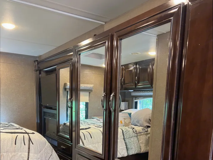 Discover comfort in this RV rental bedroom featuring dark wood mirrored cabinets, a built-in TV, and a cozy bed with laminate flooring.. Thor Quantum 2017