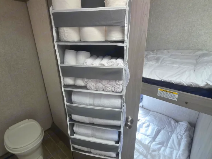 Inside an RV rental: a hanging organizer with towels and toilet paper, next to a toilet and bunk beds with white bedding.. Dutchmen Coleman Lantern LT 2022