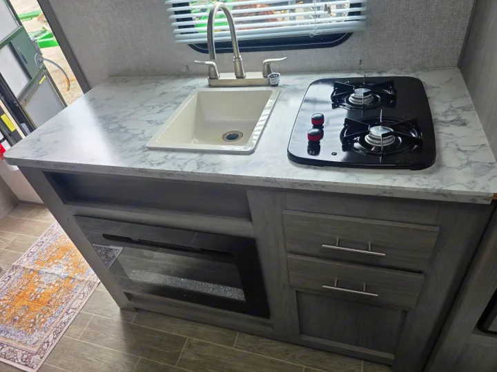 Modern RV rental kitchen features: marble-look counter, sink, two-burner stove, dark cabinets, and electric fireplace.. Dutchmen Coleman Lantern LT 2022