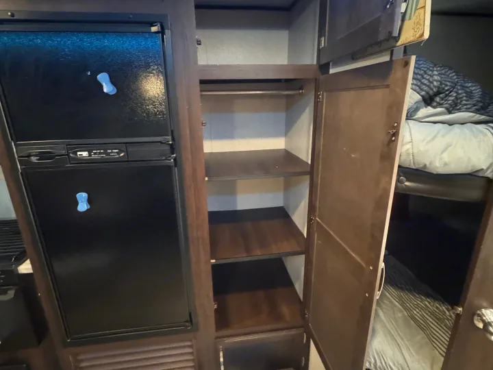 Inside an RV rental: a black refrigerator, an open wardrobe with shelves, and a glimpse of a bunk bed with striped bedding.. Jayco Jay Flight SLX 2020