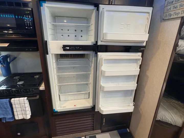 The compact kitchen of an RV rental features an open fridge/freezer, microwave, stove, and nearby bunk bed.. Jayco Jay Flight SLX 2020