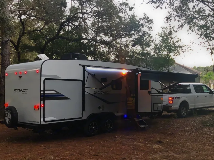 White Sonic RV rental with awning extended & lights on, hitched to a truck in a wooded campsite, ready for adventure.. Sonic Sonic Trailer 2022