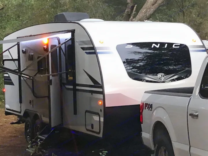 White travel trailer RV rental, partially towed by a pickup truck, with awning & lights extended at a campsite.. Sonic Sonic Trailer 2022
