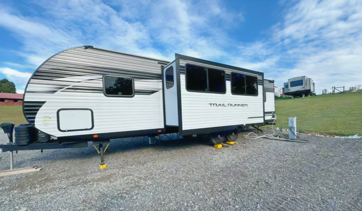 This white and grey Trail Runner travel trailer, an RV rental, is parked with its slide-out extended at a gravel campsite.. Forest River Cherokee 2022