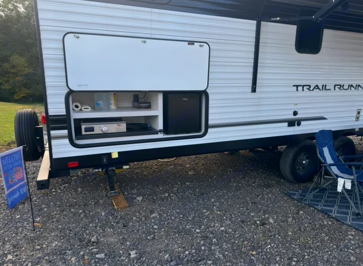 RV rental showing an open exterior kitchen with a grill, mini-fridge, and supplies. Camp chairs and a mat are on the gravel.. Forest River Cherokee 2022