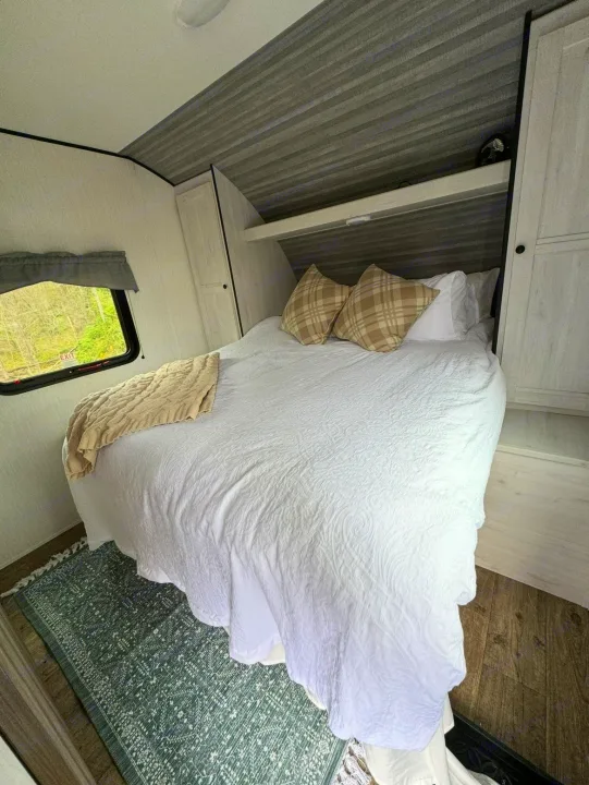Cozy RV rental bedroom with white bedding, plaid pillows, and a window looking out to green trees, featuring a soft rug.. Forest River Cherokee 2022