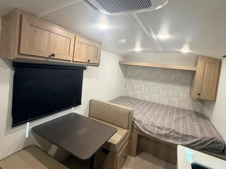 Compact RV rental interior featuring a dinette with bench seating, black roller blind, and bed with plaid bedding. Overhead cabinets provide storage in this efficient space.. Forest River Rockwood Geo Pro 17LE 2026
