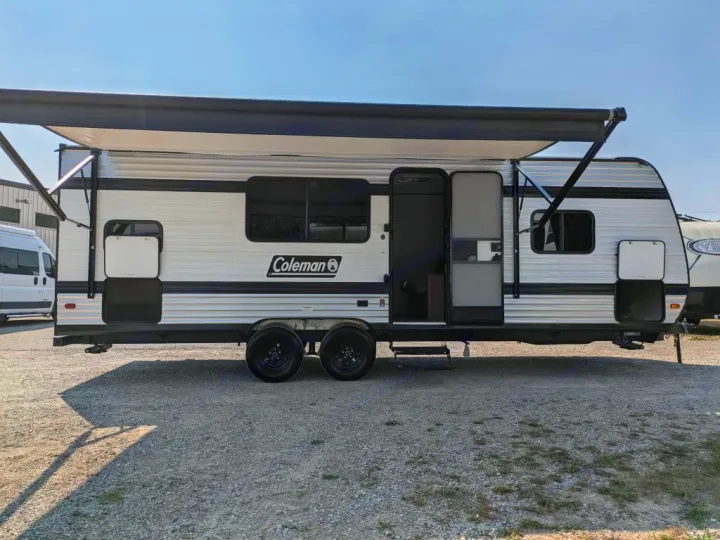 White Coleman rv rental travel trailer on gravel, awning extended. Open door reveals screen. Ideal for camping & road trips.. Keystone RV Coleman 2025