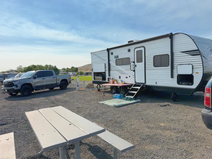 RV rental setup: A white travel trailer and grey pickup truck parked at a sunny gravel campsite with a picnic table and outdoor seating.. Jayco Jay-flight 2026