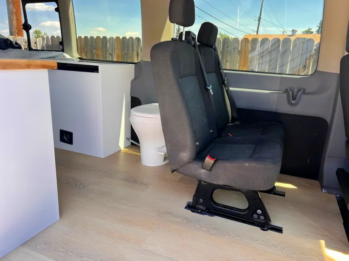 RV rental interior: two black passenger seats, a portable toilet, and light cabinetry inside a van conversion.. Ford Transit 2016