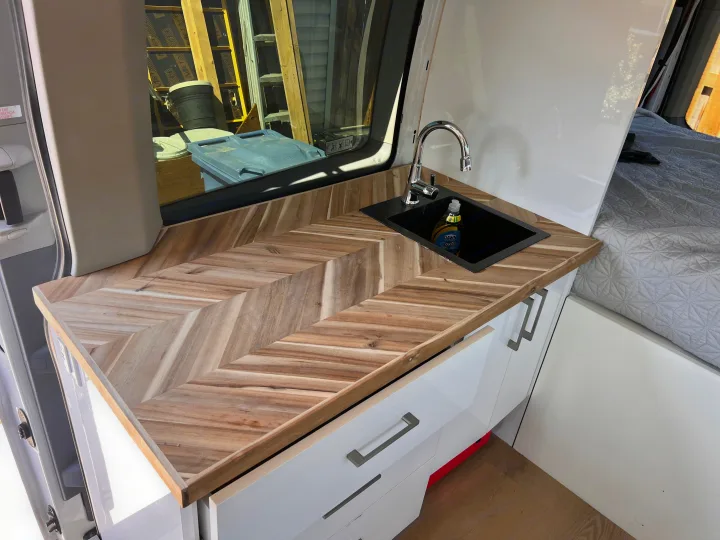 Inside an RV rental: stylish kitchen with wood herringbone counter, sink, white cabinets, and a bed.. Ford Transit 2016
