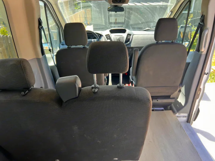 Inside an RV rental van: view from the rear of multiple rows of black fabric seats, light wood-look floor, and the dashboard.. Ford Transit 2016