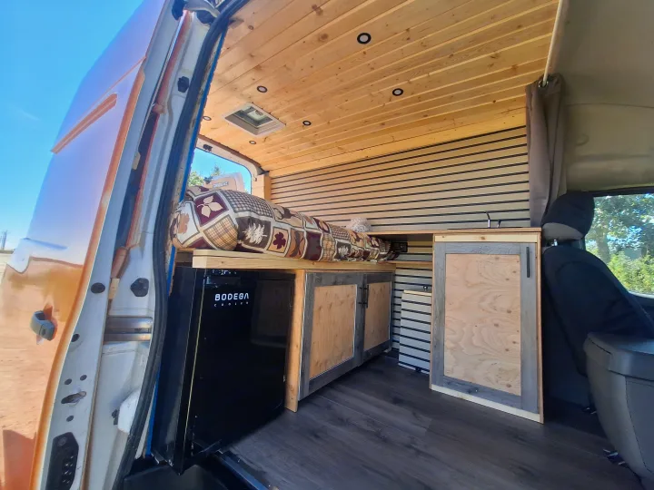 RV rental van interior: wood ceiling, bed, Bodega Cooler fridge, wood cabinetry, dark floor. Bright exterior through open door.. Glacier Campers Nissan NV 3500 2013