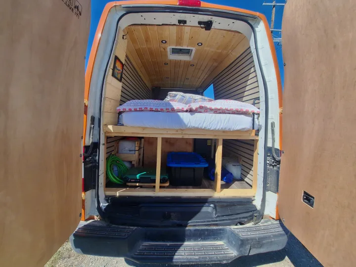 Inside an RV rental camper van, showing a bed platform, under-bed storage with gear, and wood-lined interior.. Glacier Campers Nissan NV 3500 2013