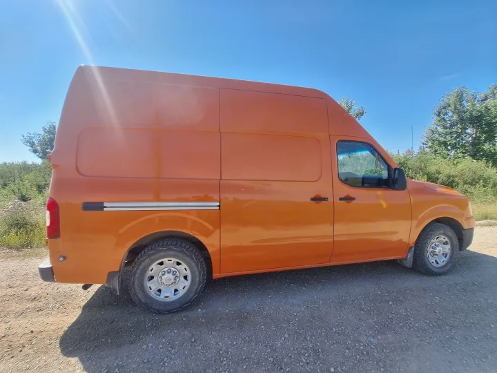 Bright orange high-top RV rental van parked on a sunny gravel path. Ideal for off-road camping and adventures.. Glacier Campers Nissan NV 3500 2013
