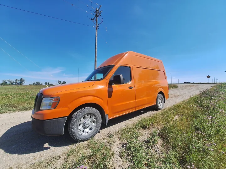 Bright orange high-top cargo van, ideal for your next RV rental road trip, parked on a dusty rural road under a clear blue sky.. Glacier Campers Nissan NV 3500 2013