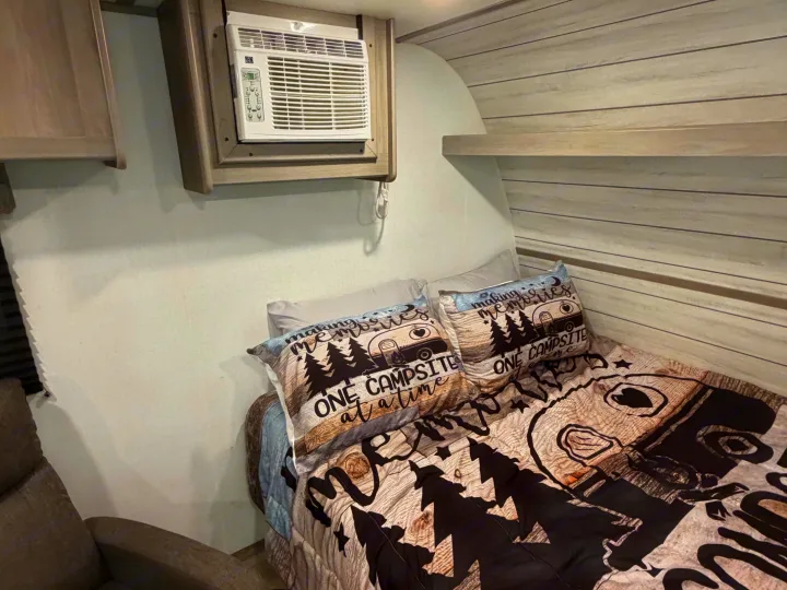 Cozy RV rental bedroom featuring a window AC unit, "making memories" themed bedding, and light wood paneling.. Dutchmen Coleman 2024