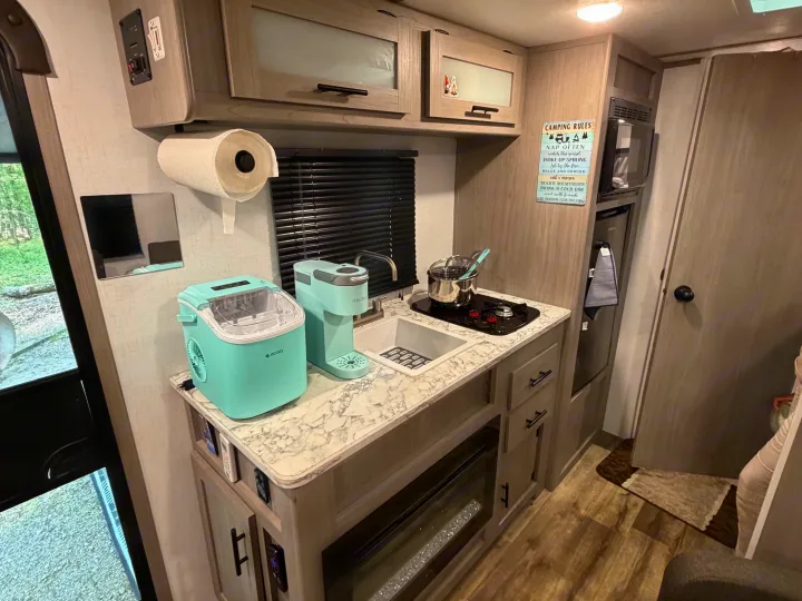 Well-equipped RV rental kitchen: teal Keurig, ice maker, sink, stovetop, and microwave with charming 'Camping Rules' sign.. Dutchmen Coleman 2024