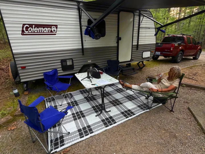 Campsite setup with an **rv rental**: Coleman travel trailer, red pickup truck, outdoor rug, and a person relaxing under the awning.. Dutchmen Coleman 2024