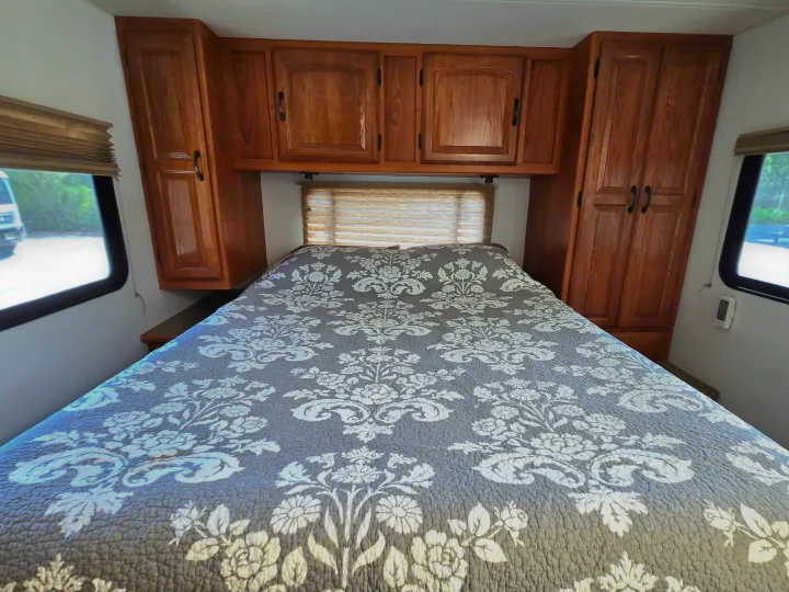 Cozy RV rental bedroom featuring a patterned bedspread, ample overhead wooden cabinets, and window blinds for privacy.. Coachmen Freedom Express 2008