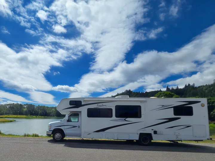 A white RV rental parked by a peaceful river, surrounded by green trees under a vibrant blue sky with fluffy white clouds.. Coachmen Freedom Express 2008