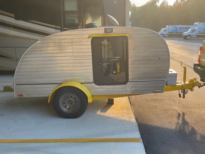 This silver teardrop camper, an RV rental option, sits ready on a concrete lot near a larger RV and moving trucks.. hand built teardrop 2016