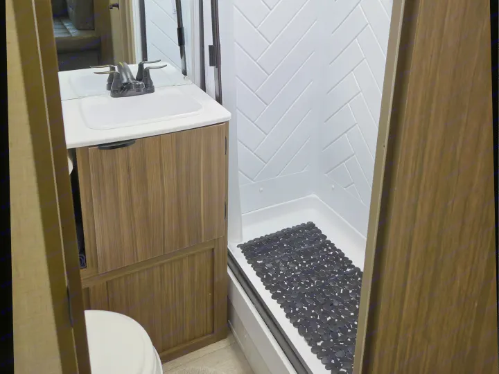 Inside an RV rental: a compact bathroom with a vanity, sink, toilet, and shower stall with a black non-slip mat.. Forest River Wildwood X-Lite 2025