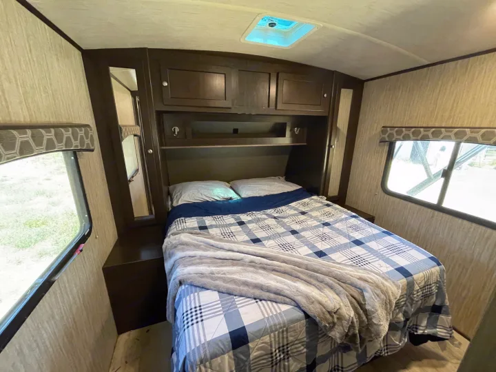 Inside a cozy RV rental bedroom: a comfy bed with plaid bedding, overhead storage, and windows offering natural light.. Cruiser RV Shadow Cruiser 2019