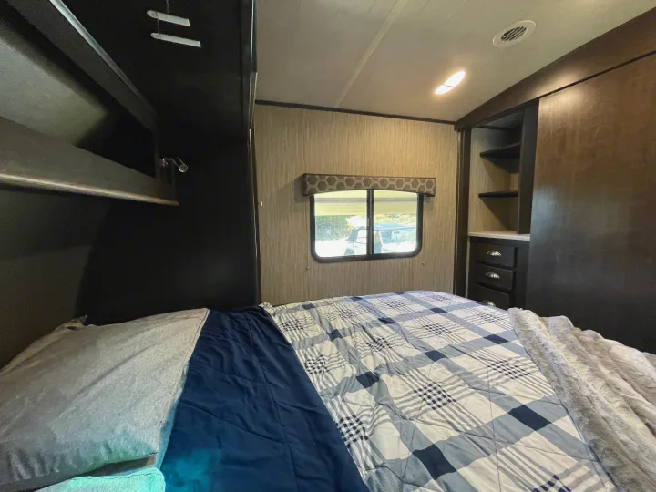Cozy RV rental bedroom featuring a comfortable bed with plaid comforter, window view, and built-in storage.. Cruiser RV Shadow Cruiser 2019