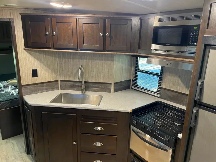 Inside an RV rental: a compact kitchen with a stainless steel sink, microwave, stove, and refrigerator, plus dark cabinets.. Cruiser RV Shadow Cruiser 2019