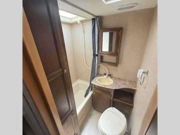 Compact RV rental bathroom with shower, toilet, and sink vanity. Features dark wood cabinetry and light, textured walls.. Coachmen Apex Nano 2018