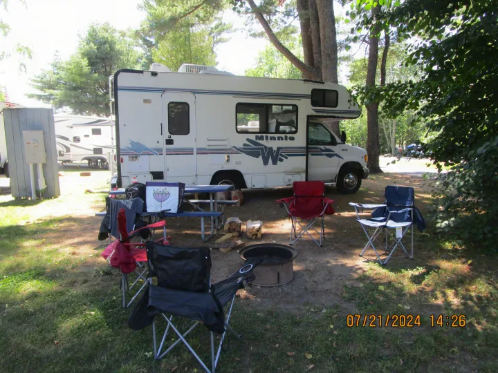 RV rental at a shaded campsite. A Winnebago Minnie, camping chairs, fire pit, and picnic table are set up.. Winnebago Minnie 1998