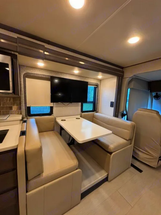 Modern rv rental interior showing a beige dinette with a table, wall-mounted TV, and an adjacent kitchen counter.. Thor Palazzo 2023