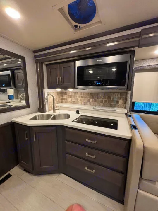 Modern RV rental kitchen showing dark cabinets, double sink, induction cooktop, microwave, and tiled backsplash.. Thor Palazzo 2023