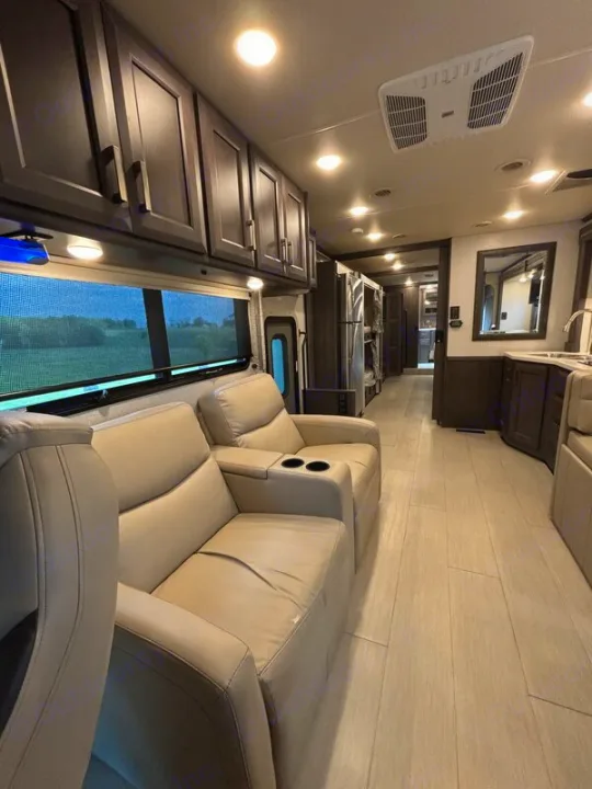 Modern RV rental interior with cream recliners, dark cabinets, light wood flooring, and a full kitchen area. Bright living space.. Thor Palazzo 2023