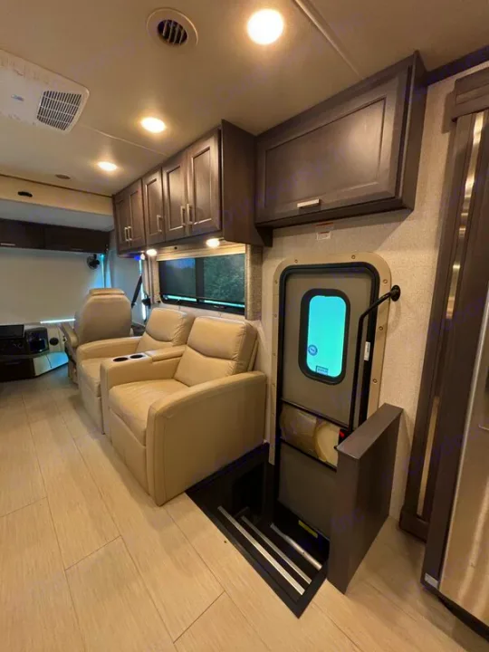 Bright RV rental living space featuring light wood-look flooring, two beige recliners, overhead cabinets, and an RV entry door with retractable steps.. Thor Palazzo 2023