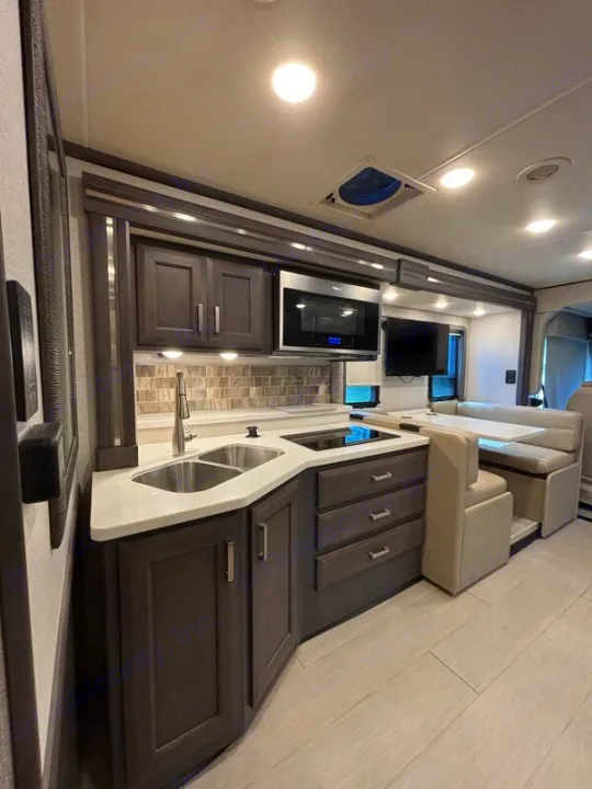 Modern RV rental kitchen with dark cabinets, light counters, sink, cooktop, microwave, and dinette area.. Thor Palazzo 2023