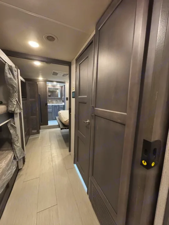Modern RV rental interior: a hallway with light flooring, bunk beds left, dark cabinets right, leading to a kitchen/bath area.. Thor Palazzo 2023