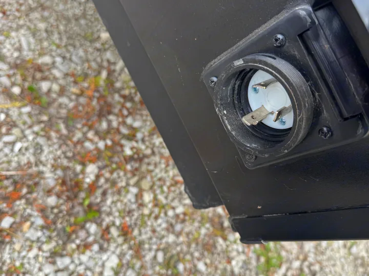 RV rental power inlet. Side view of a black electrical inlet with three metal prongs (two flat, one L-shaped), mounted on an RV over gravel.. Forest River Coachmen Clipper 2023