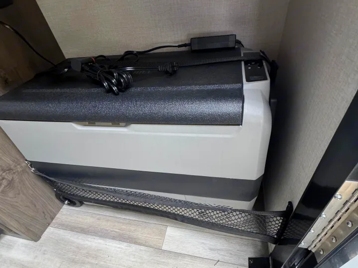 An RV rental's portable fridge, powered via a coiled cord, sits on the floor with a mesh storage net attached below it.. Forest River Coachmen Clipper 2023