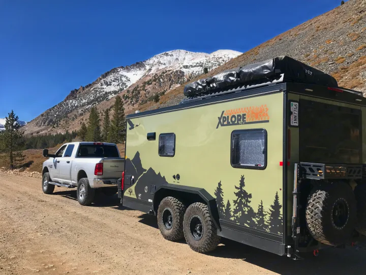 XPLORE RV rental trailer towed by a truck on a rugged mountain road, showcasing off-road adventure with snow-capped peaks.. Imperial Outdoors XploreRV 2022
